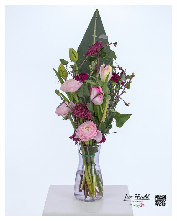 Moderner Blumenstrauß mit rosa Ranunkeln, violetten Nelken, gefärbtem Ammi und zarten Tulpen, kombiniert mit dekorativen Kirschzweigen und einem großen Strelitzienblatt als Hintergrund.