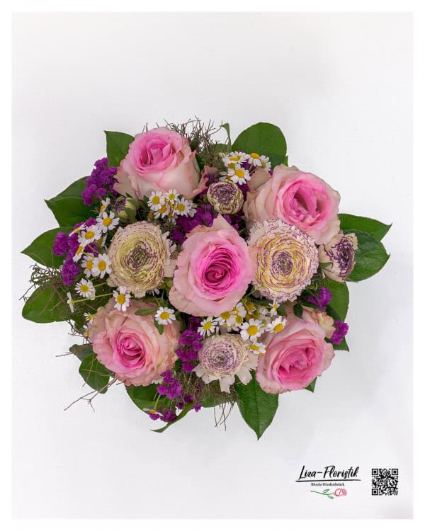 Blumenstrauß in harmonischer Rundbindung mit sieben großblütigen Ecuador-Rosen in Rosé- und Pinktönen, ergänzt durch cremefarbene Ranunkeln mit violett gezeichneter Mitte. Akzentuiert mit kleinen weißen Kamilleblüten, kräftig violettem Strandflieder und lockerem Beiwerk. Das frische Grün bildet einen natürlichen Rahmen um das blütenreiche Zentrum.